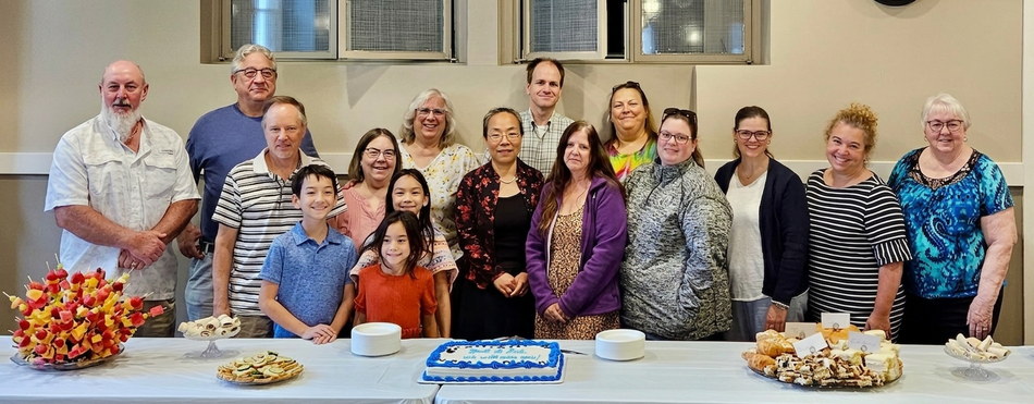 Members of the Trinity Evangelical Lutheran congregation, adults and children, standing together behind a reception table set with a decorated cake, fruit skewers, and sandwich trays.